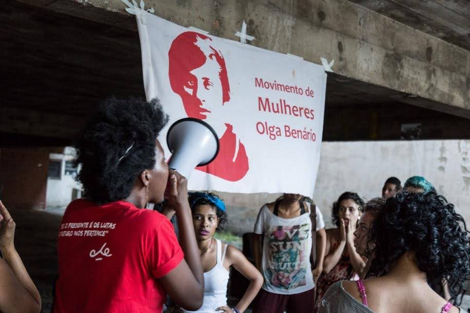 Movimento Olga Benario se prepara para o I Encontro das Ocupações, nos dias 30 e 31 de maio, em São Paulo. Na foto, uma militante com um mega fone, no fundo da imagem há a bandeira do Movimento de Mulheres Olga Benario.