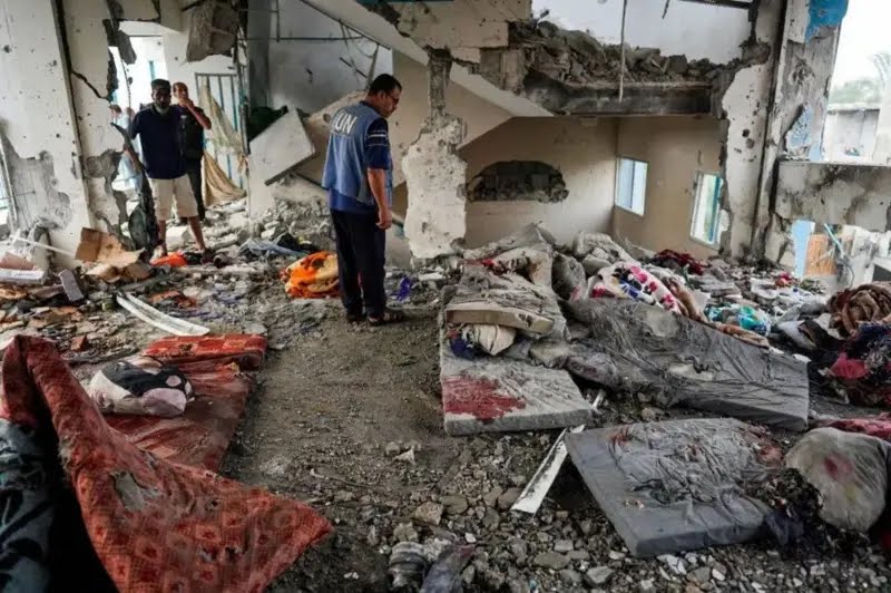Colchões espalhados em prédio destruído em escola da ONU bombardeada por israel. No centro, um funcionário das Nações Unidas observa a destruição.
