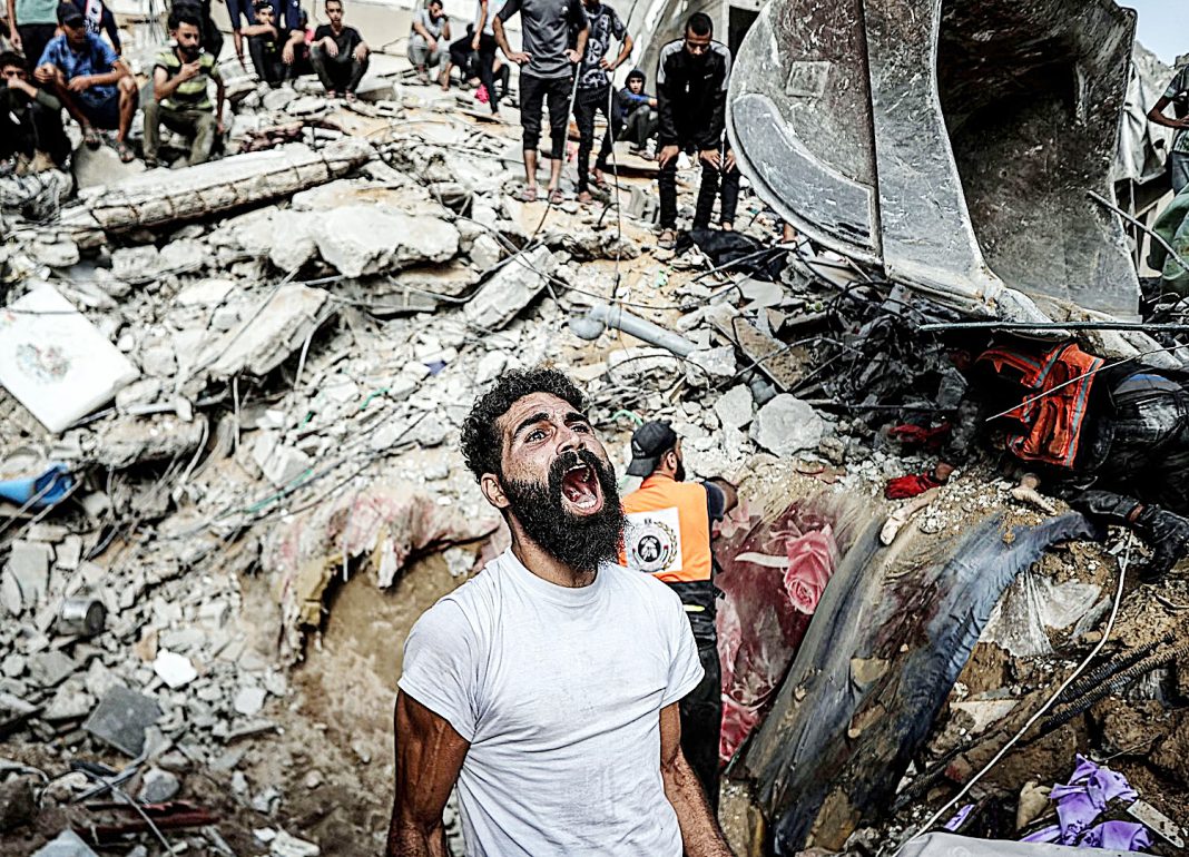 Palestinos observam escombros de campo de refugiados bombardeado no genocídio de Gaza, ao centro um homem vestido de branco grita de desespero diante da destruição.