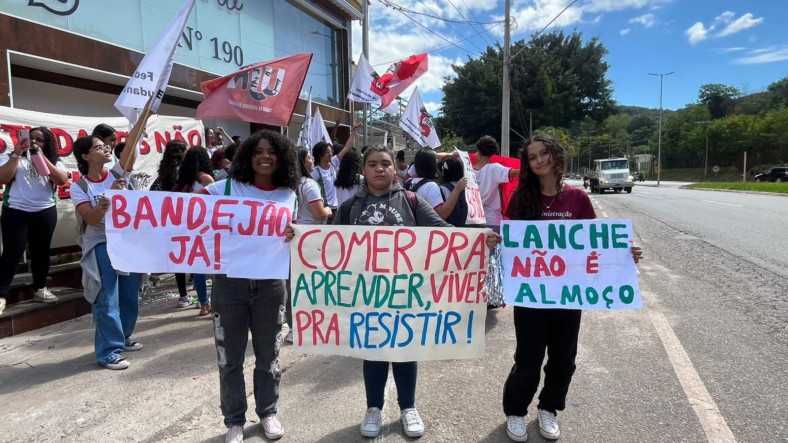 Três estudantes seguram cartazes com frases como “Bandejão já!”, “Comer pra aprender, viver pra resistir!” e “Lanche não é almoço” durante protesto por alimentação escolar digna na Grande BH.