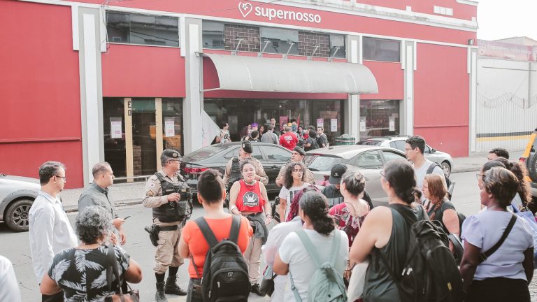 Fotografia colorida mostra um grupo de manifestantes e trabalhadores reunidos em frente ao supermercado Supernosso, de fachada vermelha. Dois policiais militares acompanham a movimentação. No centro, uma jovem com camiseta vermelha do MLC lidera a conversa.
