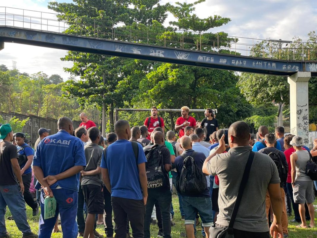 Fotografia colorida em plano médio de uma assembleia de trabalhadores ao ar livre. Em primeiro plano, dezenas de operários estão de costas, ouvindo atentamente; um deles veste um uniforme azul com o logotipo 