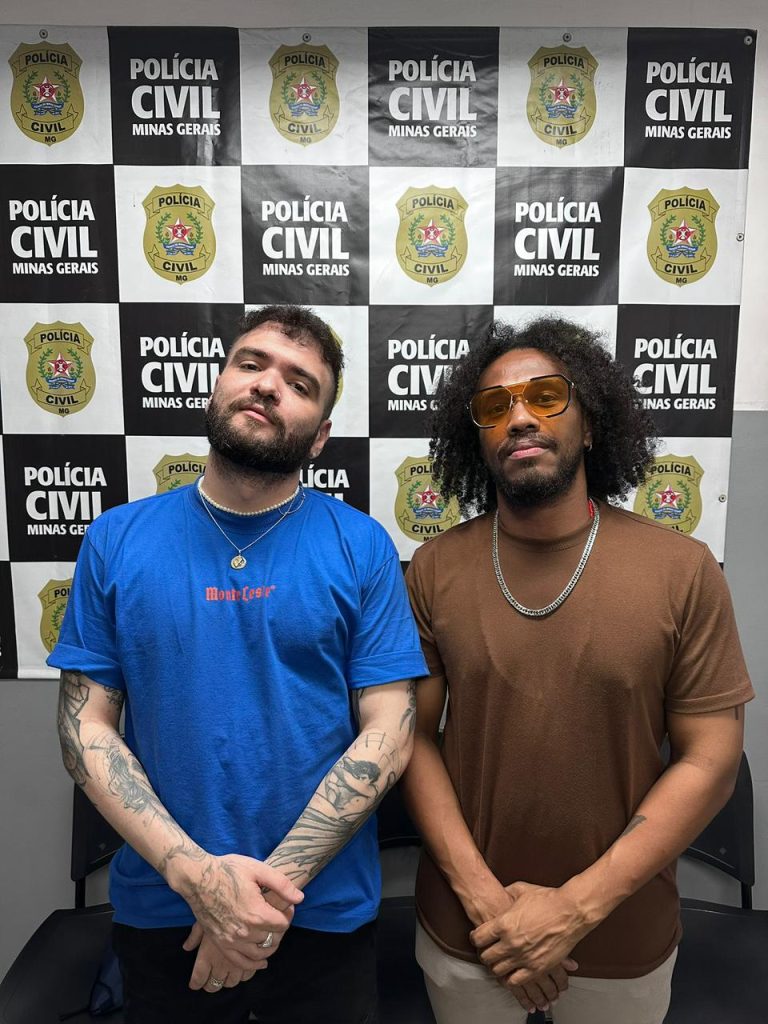 Fotografia dos humoristas Tiago Santineli e Luis Titoin em pé, um ao lado do outro, dentro de uma delegacia. Ao fundo, há um painel xadrez em preto e branco com o brasão e o nome da Polícia Civil de Minas Gerais. Tiago, à esquerda, veste camiseta azul e exibe tatuagens nos braços. Luis Titoin, à direita, é um homem negro com cabelos estilo afro, veste camiseta marrom e óculos de lentes laranjas.