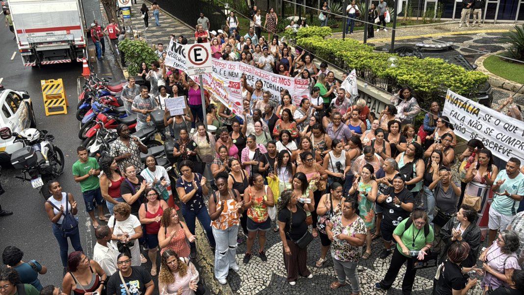 Fotografia em ângulo alto mostra uma multidão de trabalhadoras terceirizadas da educação, em sua maioria mulheres negras, reunidas em protesto na calçada e em parte da rua em frente ao TRT, em Belo Horizonte. Elas batem palmas e seguram faixas com mensagens contra a prefeitura e a empresa MGS. O chão está molhado pela chuva e há motocicletas estacionadas ao lado da concentração.