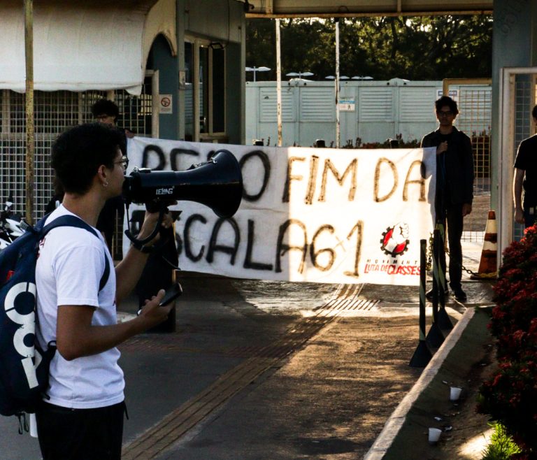 Fotografia em plano médio de um militante jovem, de costas, segurando um megafone preto e falando em direção à entrada de uma empresa. Ao fundo, dois jovens seguram uma grande faixa branca onde se lê, em letras pretas garrafais: "PELO FIM DA ESCALA 6x1". No canto direito da faixa, aparece a logomarca do Movimento Luta de Classes (MLC) com um punho cerrado. A cena ocorre ao ar livre, durante o dia, sob a luz do sol.