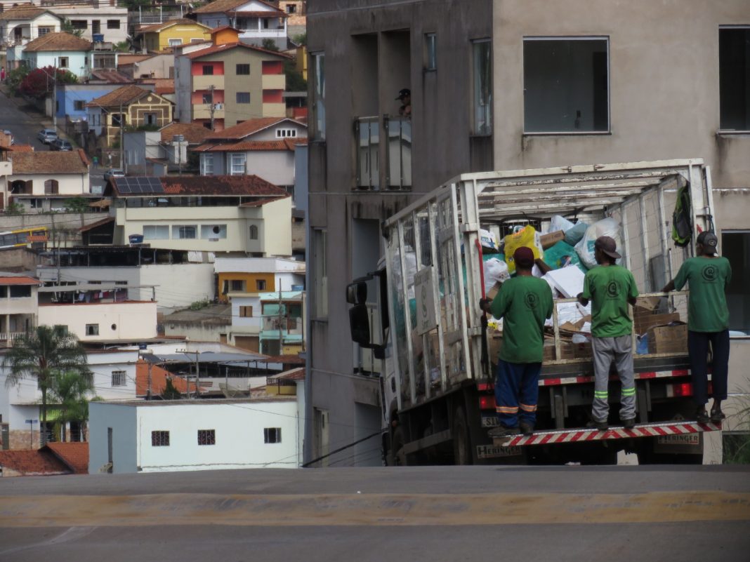 Três trabalhadores da limpeza urbana, vestindo uniformes verdes e bonés, estão em pé no estribo traseiro de um caminhão de lixo branco carregado. O caminhão sobe uma rua íngreme em Itabirito, Minas Gerais. Ao fundo, vê-se uma paisagem urbana composta por diversas casas construídas em morros.