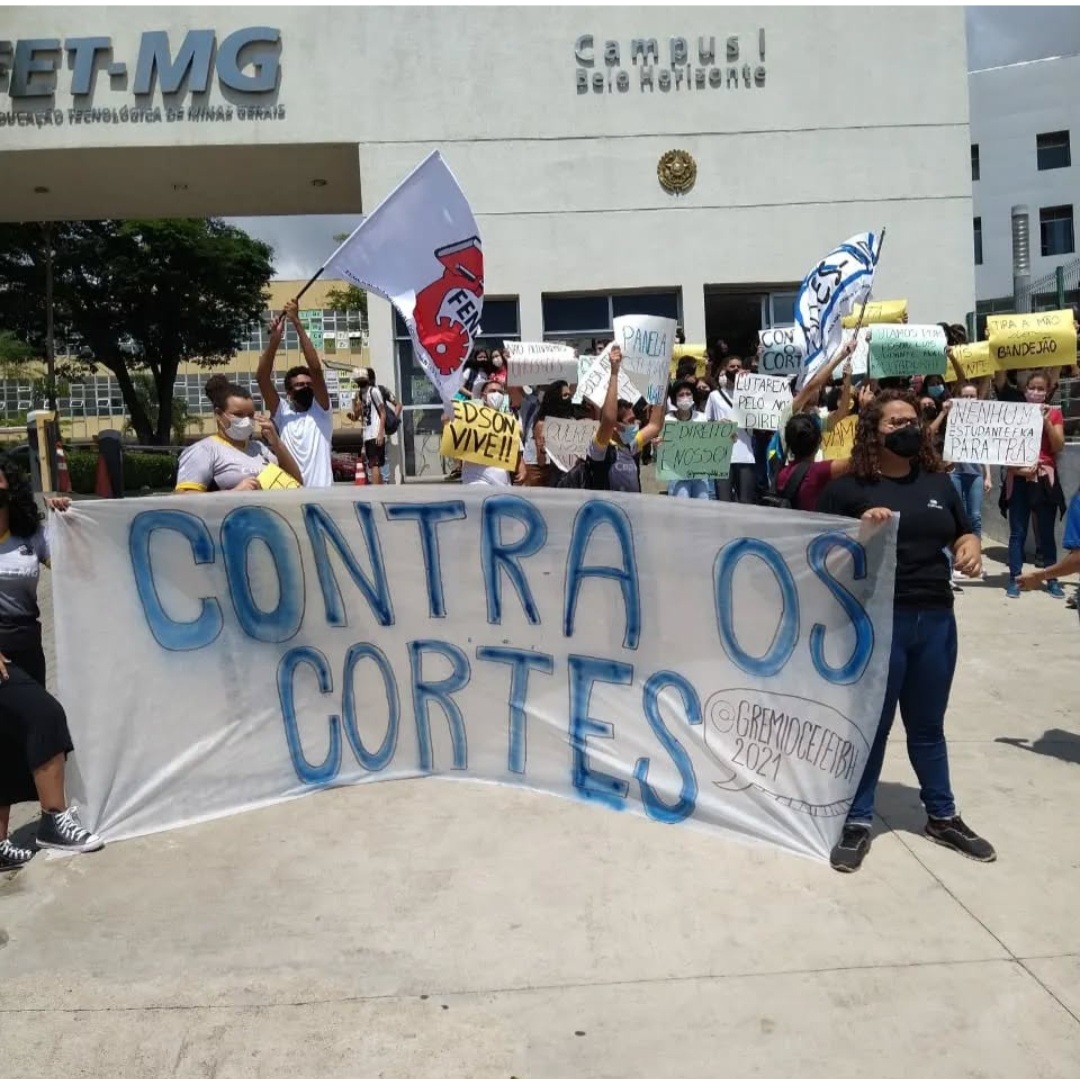 Estudantes em frente ao campus do CEFET-MG seguram uma grande faixa branca onde se lê, em letras azuis, "CONTRA OS CORTES". Ao fundo, jovens seguram bandeiras da FENET e cartazes com dizeres como "EDSON VIVE!!" e "NENHUM ESTUDANTE PARA TRÁS".