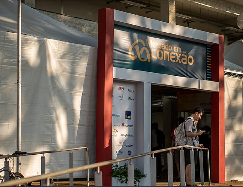Fotografia da entrada do evento “Mercado em Conexão” na Escola de Engenharia da UFMG. A estrutura conta com um pórtico central exibindo o nome do evento e, na lateral, um painel branco com logomarcas de diversas empresas patrocinadoras, entre elas Itaú, Ambev e Embraer. À direita, um estudante de mochila caminha em frente à entrada.